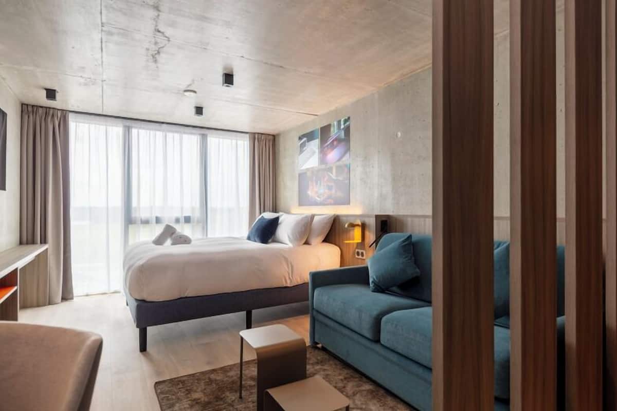 A modern hotel room with a large bed, a sofa, a small table, and a large window.