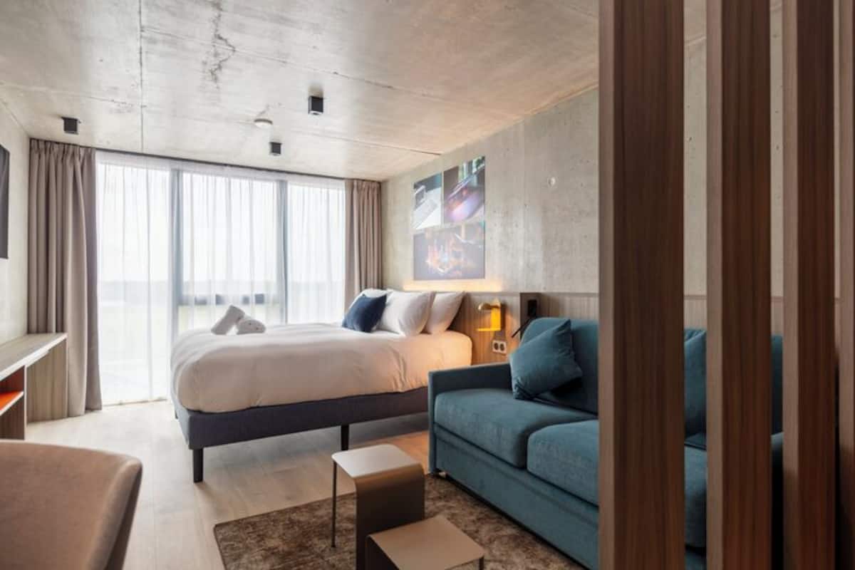 Ein modernes Hotelzimmer mit einem großen Bett, einer Sitzecke, einem kleinen Tisch und einem großen Fenster.