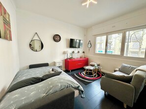 Un dormitorio con cama, un aparador rojo, un televisor y una ventana con vistas a edificios.