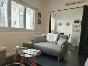 A living room with a grey sofa, a glass coffee table, a small side table, a refrigerator, and a window with a view of buildings outside.