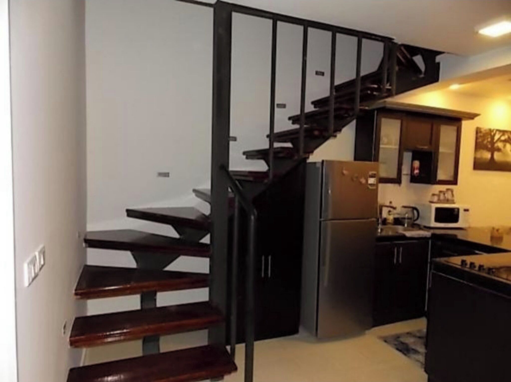 Une cuisine compacte équipée d’une cuisinière intégrée, d’un réfrigérateur et d’un four à micro-ondes. Un escalier en bois avec une rampe noire mène à un palier.