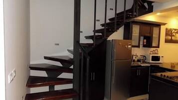 Une cuisine compacte équipée d’une cuisinière intégrée, d’un réfrigérateur et d’un four à micro-ondes. Un escalier en bois avec une rampe noire mène à un palier.