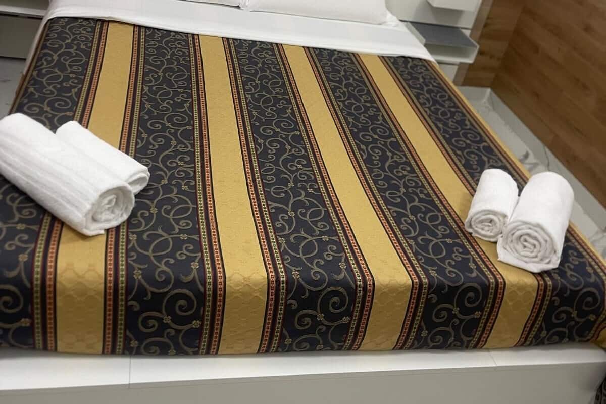 A neatly made bed with a patterned bedspread, two rolled towels, and a wooden headboard.
