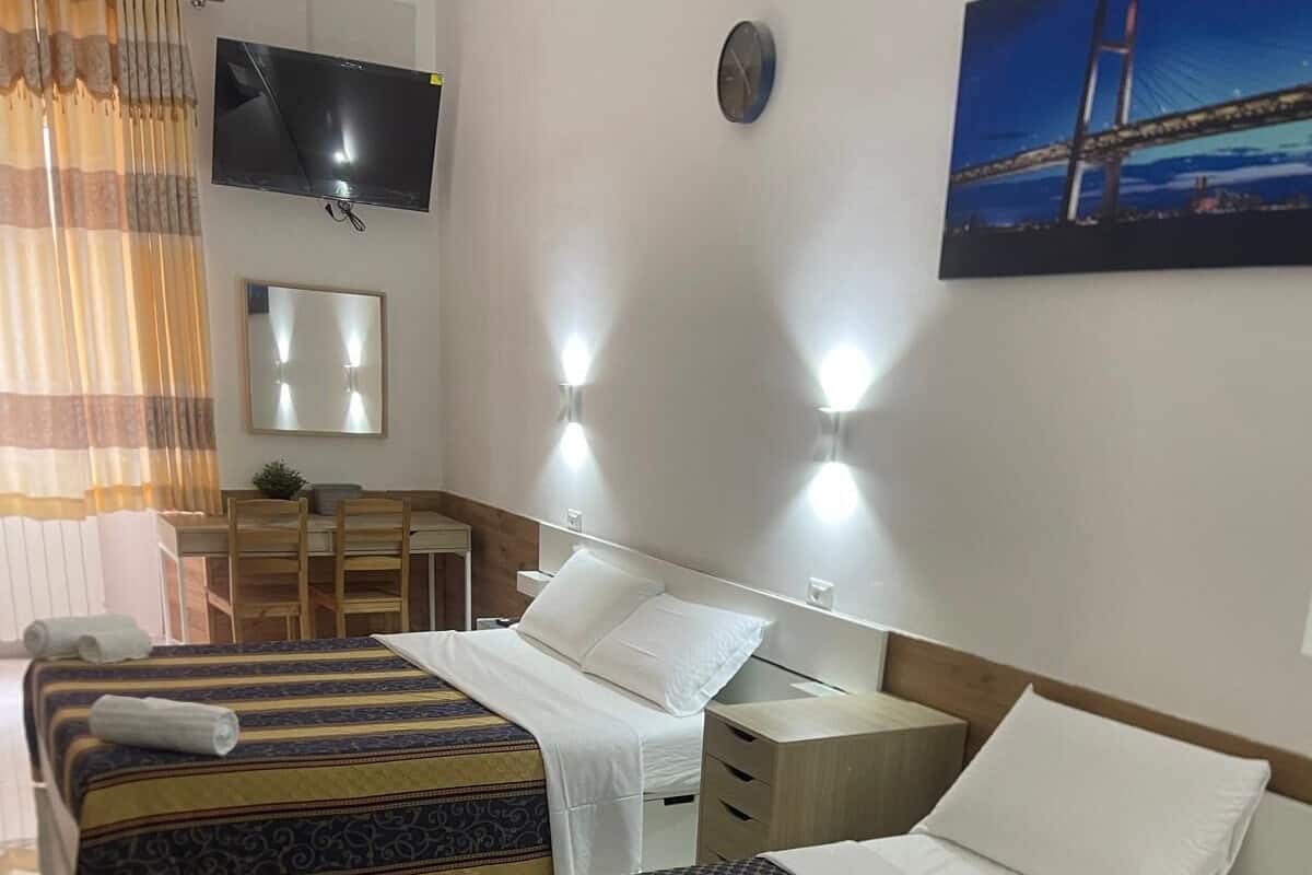 A hotel room with a bed, bedside tables, a TV, a clock, and a painting on the wall.