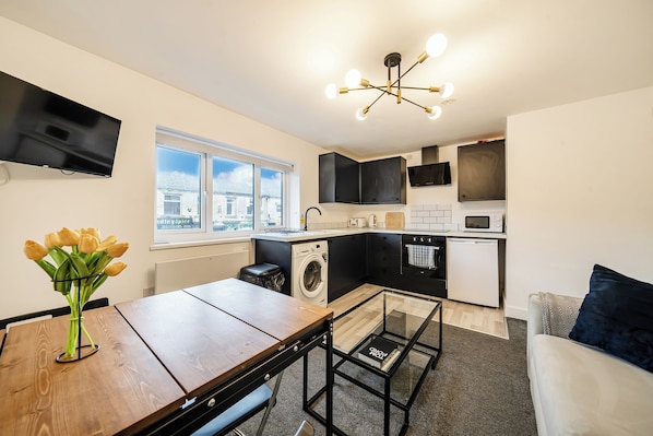 A modern kitchen with a washing machine, a dining area with a table and chairs, a sofa, and a TV mounted on the wall.