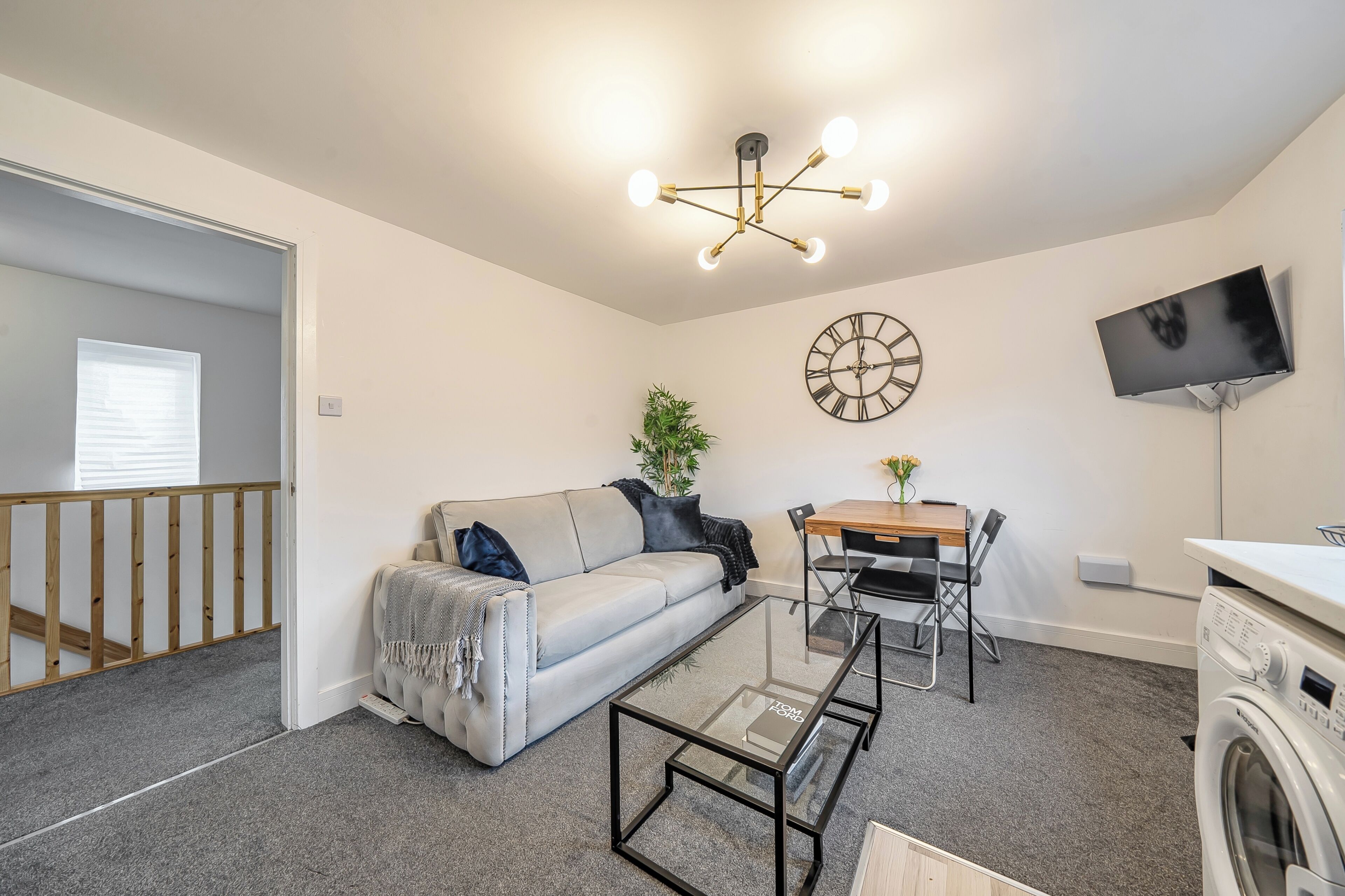 A compact living space with a sofa, a glass coffee table, a dining table with chairs, a washing machine, a TV mounted on the wall, and a clock.
