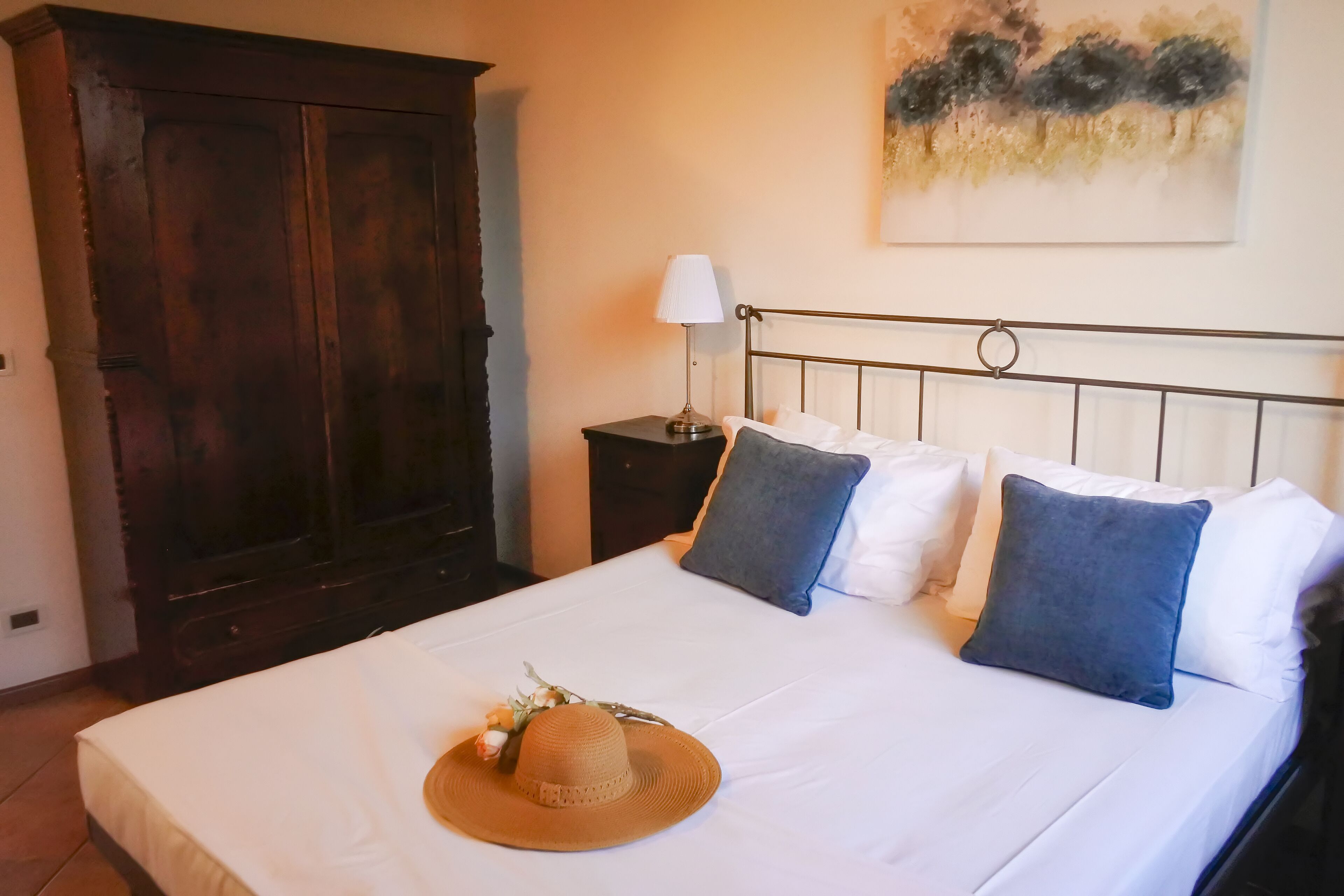 Un lit bien fait, orné d’un chapeau et de fleurs, une armoire en bois sombre, une table de chevet avec une lampe et un tableau accroché au mur.
