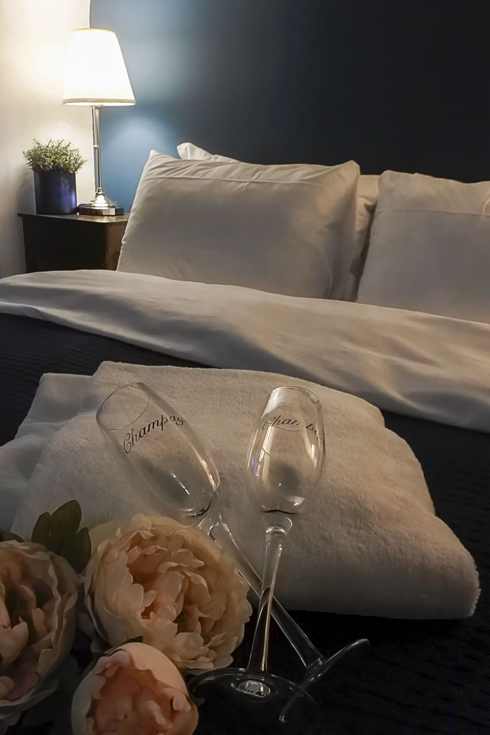 A neatly made bed with white linens, a bedside lamp, and decorative peonies.
