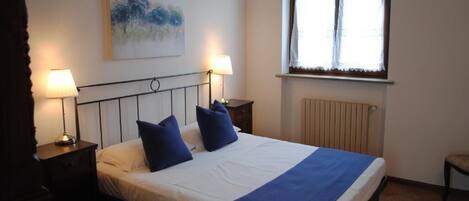 A bedroom with a bed, two bedside tables with lamps, a radiator, and a window with curtains.