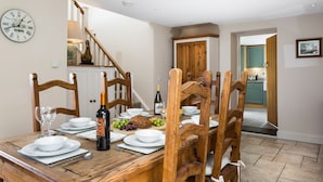Un comedor con una mesa de madera dispuesta para una comida, un reloj en la pared y una escalera que lleva a otra habitación.