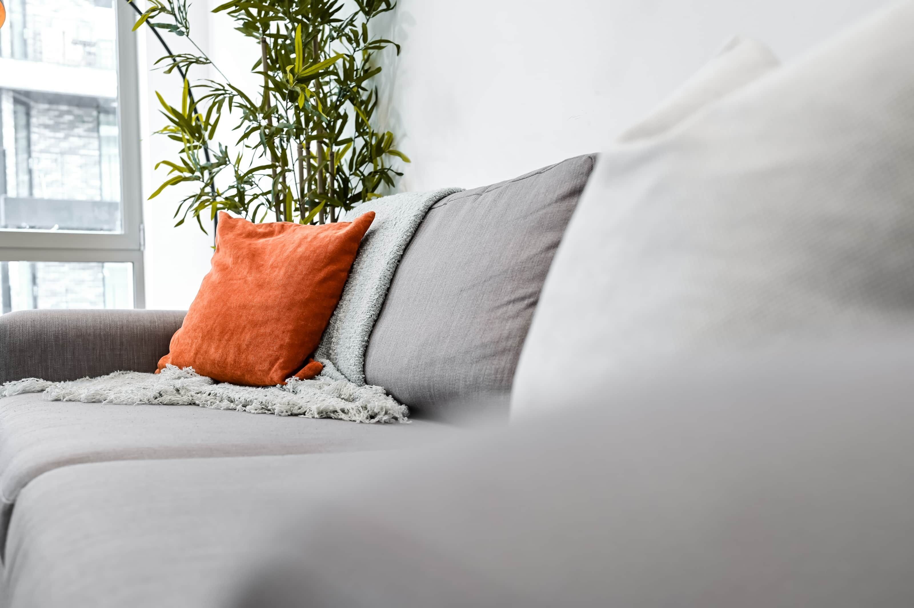Ein graues Sofa mit einer weißen Decke und einem orangefarbenen Kissen.