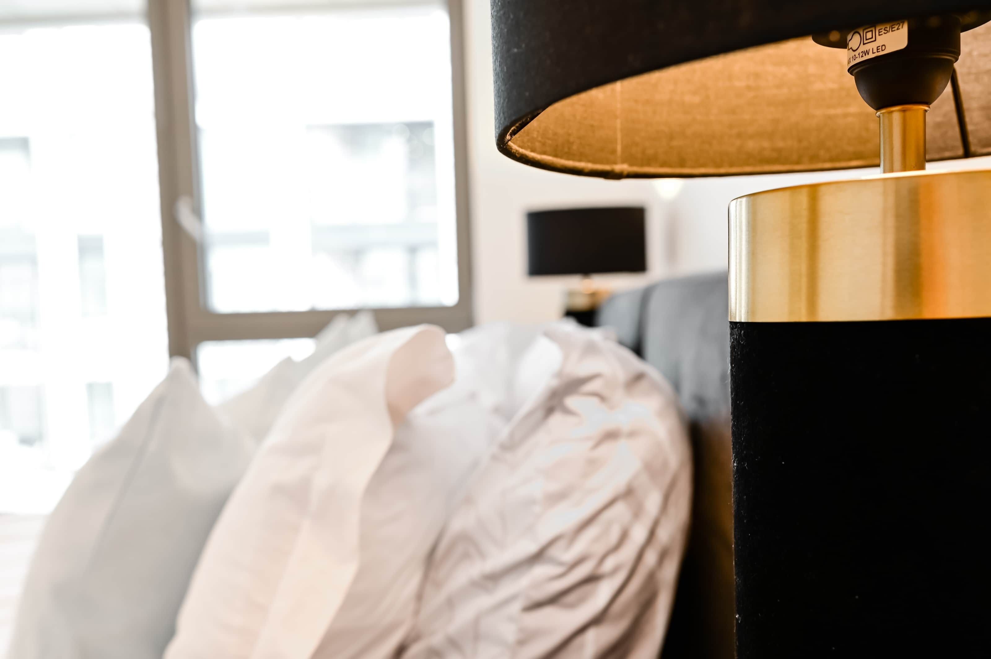 A close-up of a lamp with a black shade and a gold base, positioned next to a bed with white pillows.