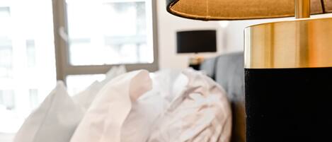 A close-up of a lamp with a black shade and a gold base, positioned next to a bed with white pillows.