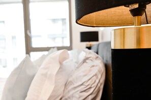 Ein Nahblick auf eine Lampe mit schwarz verziertem Schirm und goldenem Fuß, platziert neben einem Bett mit weißen Kissen.
