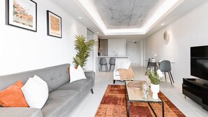 A modern living room with a grey sofa, a coffee table, and a TV. There is a dining area with a table and chairs, and a kitchen in the background.