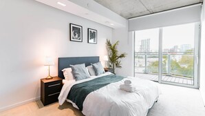 A modern bedroom with a large bed, a nightstand, a lamp, and a view of the city through large windows.