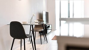 A modern dining area with a glass table, two chairs, a television, and a window with blinds.