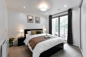 A modern hotel room with a large bed, two bedside lamps, a window with curtains, and a radiator. - The Cosmo - Two bed - Canary Wharf (London)