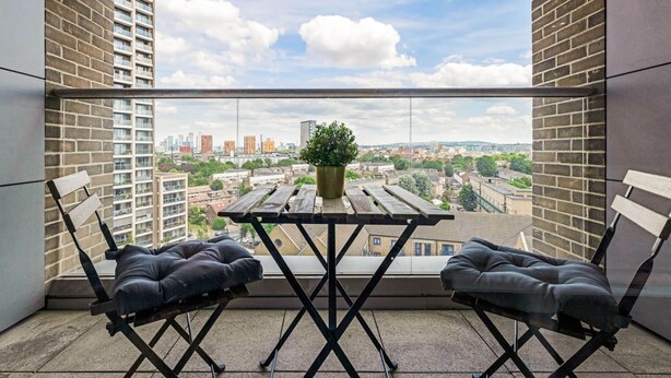 A balcony with a table and two cushioned chairs, a potted plant, and a city view. - The Ferre - Canary Wharf (London)