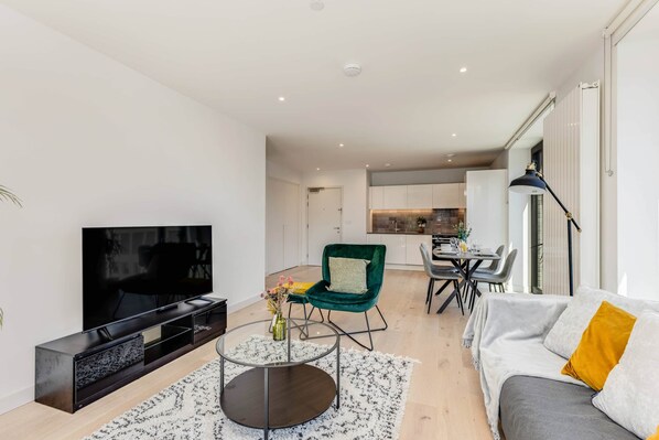A modern living room with a flat-screen TV, a green chair, a grey sofa, and a dining area in the background.
