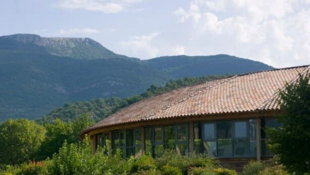 Un edifici amb teulada de rajola i grans finestres, envoltat de vegetació i muntanyes al fons.