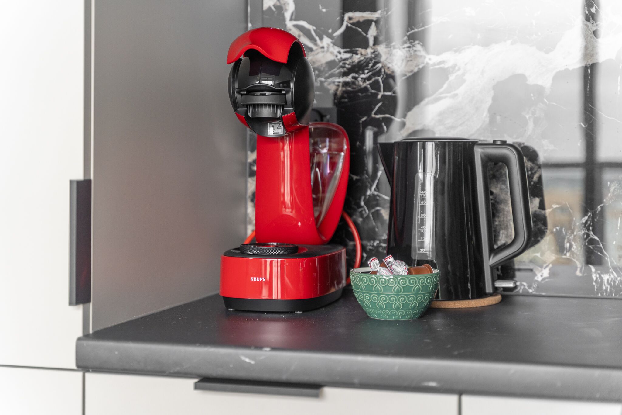 Eine rote Kaffeemaschine steht auf einer Küchenarbeitsplatte, zusammen mit einer grünen Schüssel und einer schwarzen Tasse.