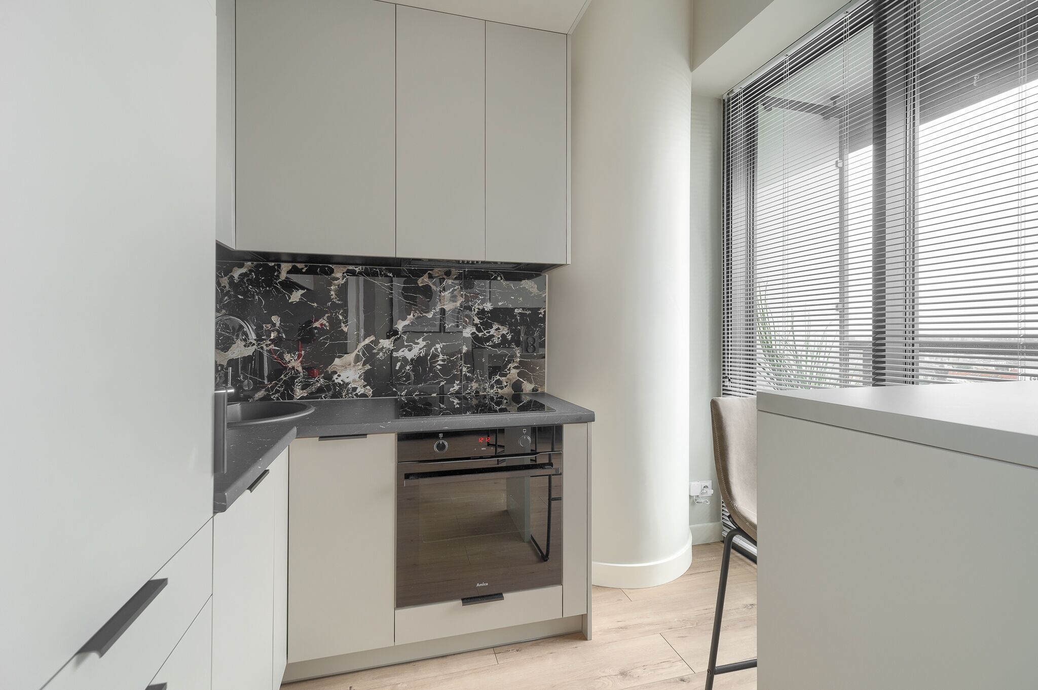 A modern kitchen with a black marble backsplash, a built-in oven, and a window with blinds.