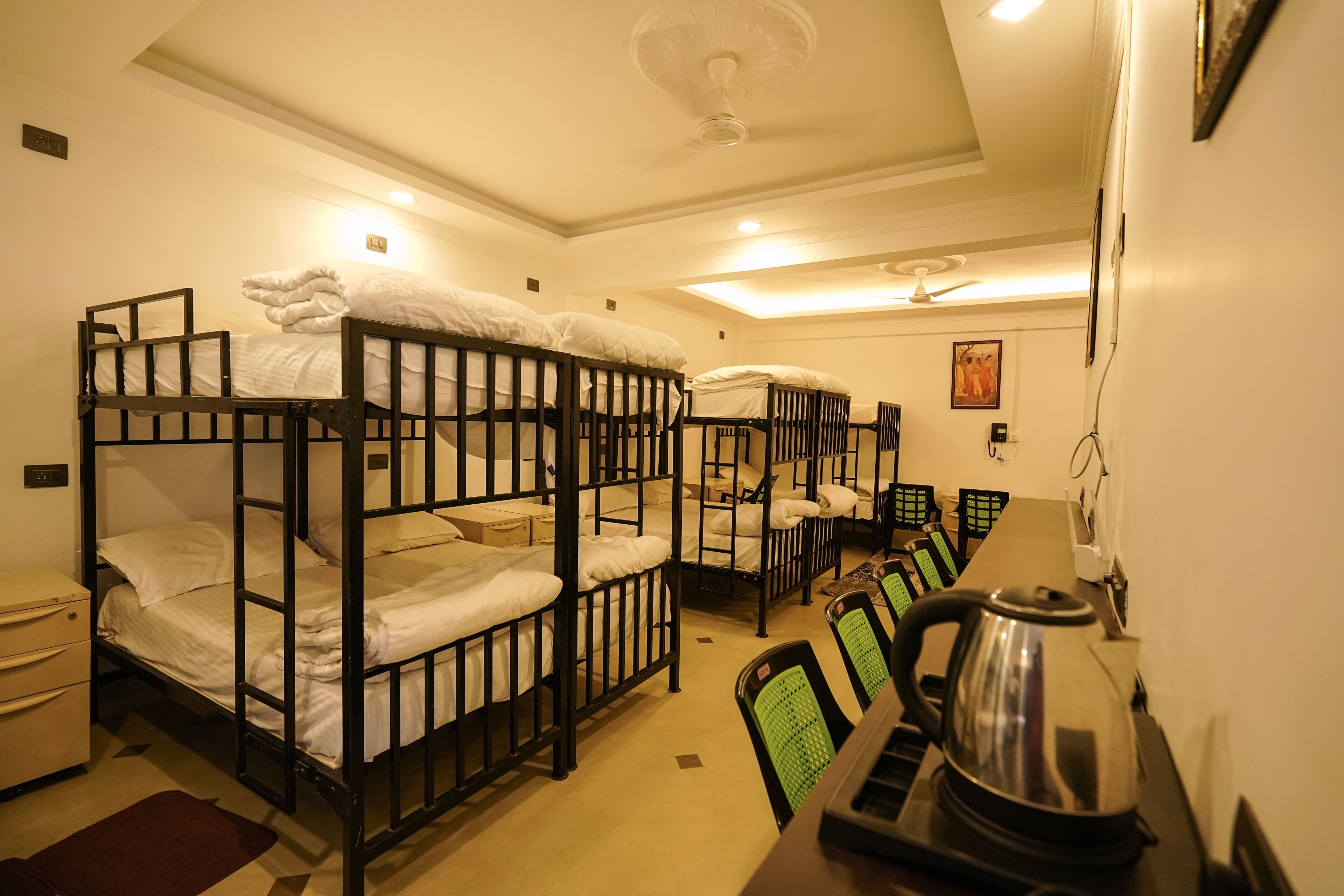 Ein Schlafsaalzimmer mit Etagenbetten, einem Schreibtisch, Stühlen, einem Wasserkocher und einem Deckenventilator.