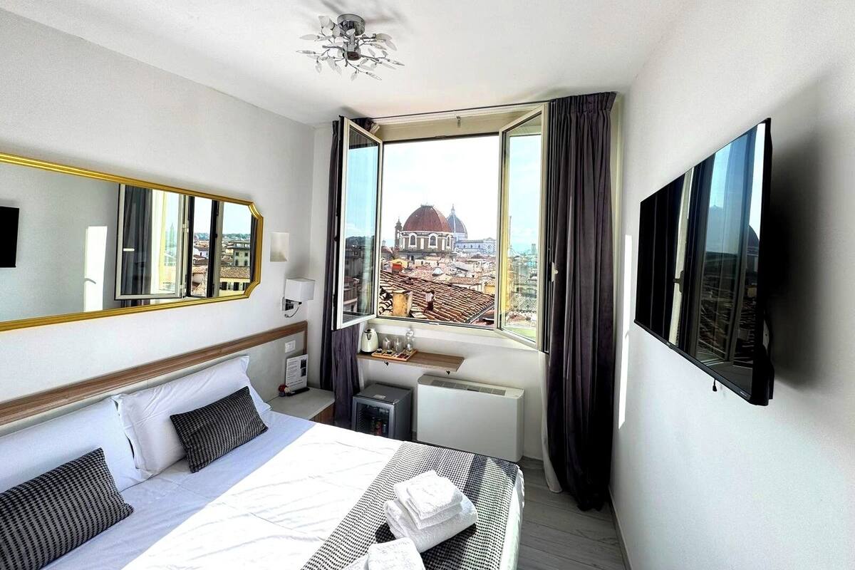 Habitación de hotel con cama, escritorio, silla, espejo, televisor y ventana con vista a la ciudad.