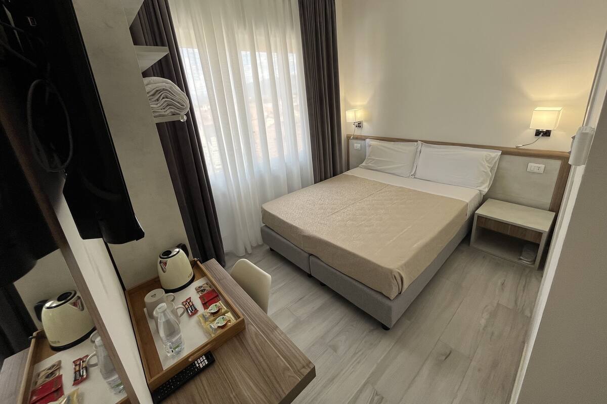Habitación de hotel con cama, mesita de noche y escritorio con hervidor, cafetera y bandeja con artículos de aseo.