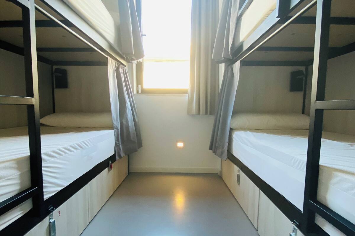 A narrow corridor with bunk beds on either side, a window at the end, and a light fixture on the wall.