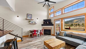 A spacious living room with a fireplace, a large window with a mountain view, a ceiling fan, and a dining area with red chairs. - Luxury Condo w/ 2 Private Balconies, Stunning Mountain Views & Natural Light! (Dillon)