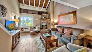 A cozy living room with a stone fireplace, comfortable seating, a wooden coffee table, and a large window offering a view of trees. - Lakeland Village #310 Modern 1 bedroom+loft (South Lake Tahoe)