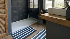 A modern bathroom with a glass shower, dark tiles, a wooden vanity, and a large window. - Chambre D'hôtes de Charme (Bernay)