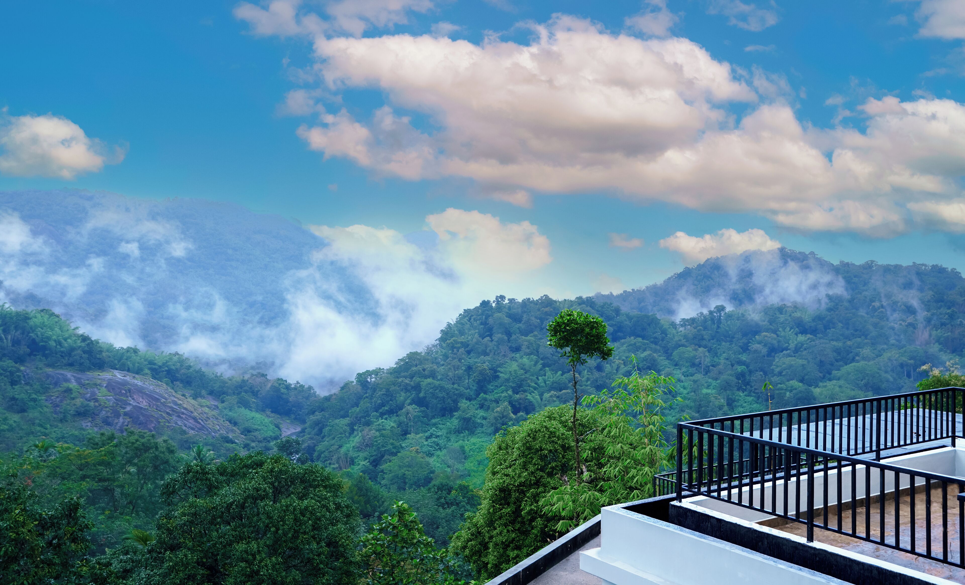 A view of a lush green valley with a mountain range in the background and a balcony with a railing.