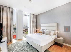 A modern hotel room with a large bed, a bedside table with a lamp, and a view of the city through large windows.