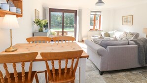 Un salón acogedor con un sofá gris, una mesa de comedor de madera y sillas. Hay una ventana con cortinas, un radiador y una lámpara.