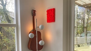 A room with a black leather sofa, a small white table with a black vase, a red abstract art piece, and a wooden guitar with a light fixture. - Prachtig Designhuis, Rustige Ligging, Zwembad, Speeltuin, Fietsen in de Duinen (De Cocksdorp)
