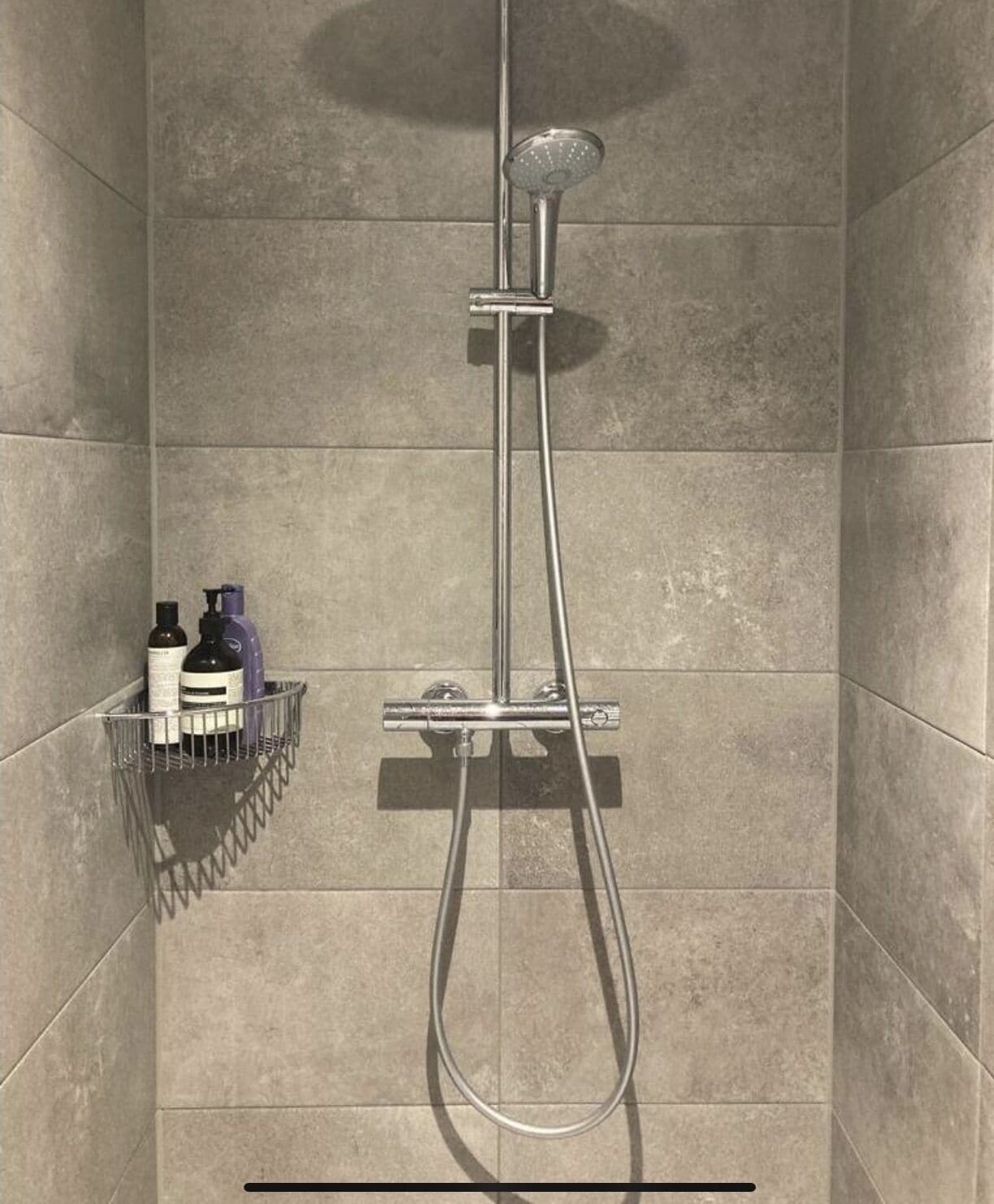 A shower with a handheld showerhead and a wall-mounted faucet, with a soap dispenser and a bottle on a shelf below.