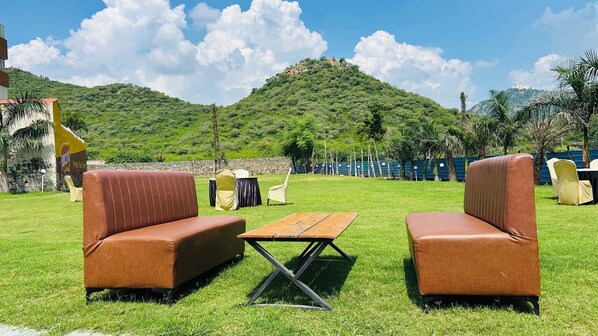 An outdoor seating area with two brown cushioned sofas and a wooden table, set against a backdrop of a grassy field and a hill with a building on top. - Raha Resort (Udaipur)