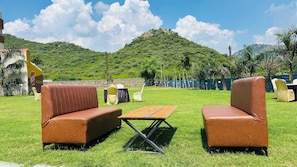 An outdoor seating area with two brown cushioned sofas and a wooden table, set against a backdrop of a grassy field and a hill with a building on top. - Raha Resort (Udaipur)