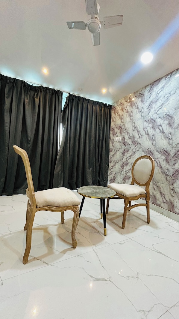 A room with two wooden chairs, a small round table, a ceiling fan, black curtains, and a marble-patterned wall.