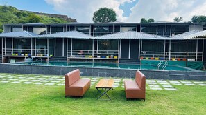 A pool area with a lounge chair and table, a swimming pool, and a building with large windows and a covered roof. - Raha Resort (Udaipur)
