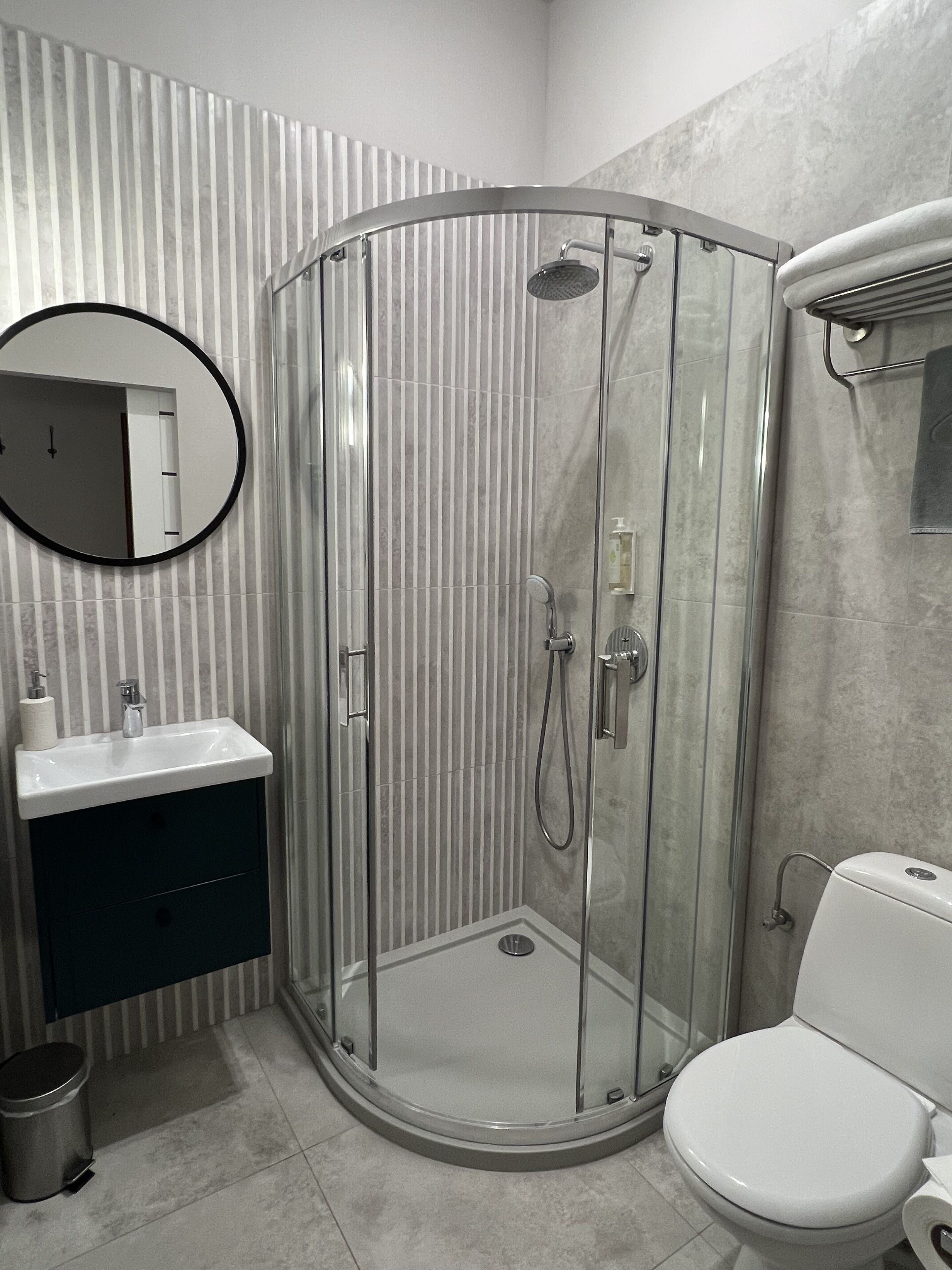 A modern bathroom with a glass-enclosed shower, a round mirror, a sink with a dark cabinet, and a toilet.