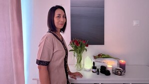 Una mujer parada en una habitación con una mesa blanca que muestra un jarrón de tulipanes rojos, una vela, una botella y una pequeña figurita.