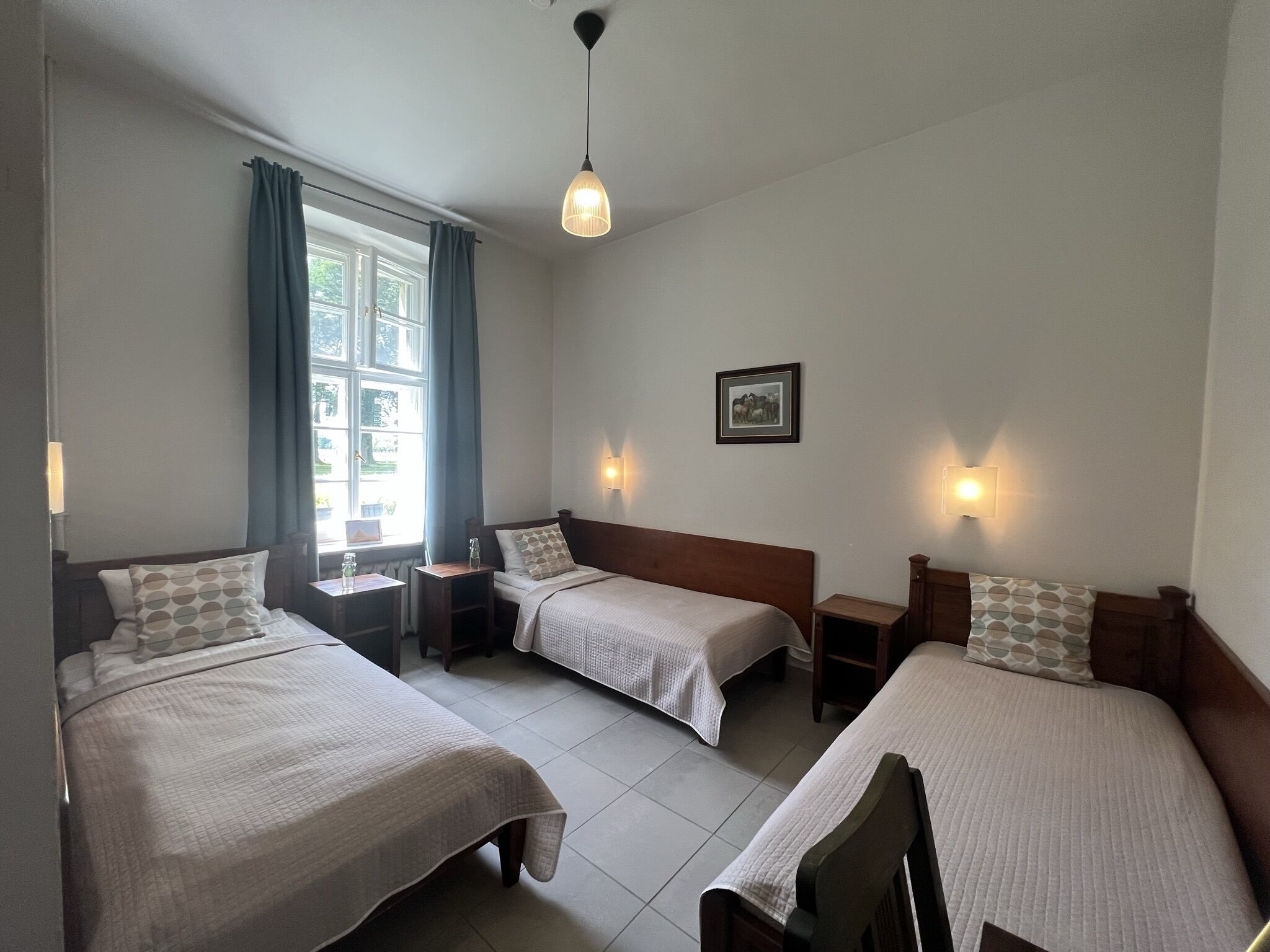 A hotel room with three single beds, a window with curtains, a wall-mounted picture, and a pendant light.