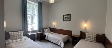 A hotel room with three single beds, a window with curtains, a wall-mounted picture, and a pendant light.