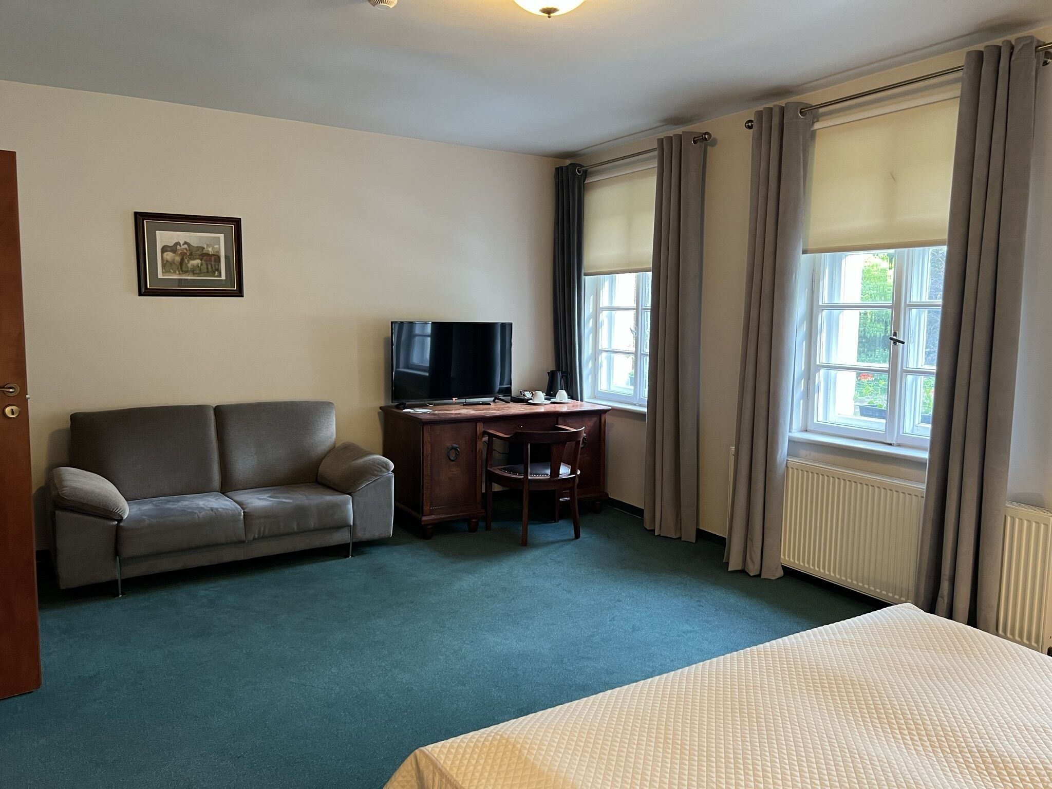 Habitación de hotel con cama, sofá, escritorio con televisor y ventanas amplias con cortinas.