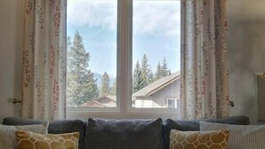 Una sala de estar con un sofá gris, dos cojines estampados y una ventana con cortinas.