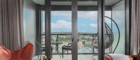 A modern balcony with a glass door, a cushioned chair, a small table with a plant, and a view of the city and sea.